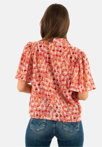 Donna con lunghi capelli castani che indossa una blusa a maniche corte floreale arancione e rossa e jeans blu, vista da dietro.