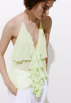 Donna con top verde chiaro a collo halter con dettagli arricciati e pantaloni bianchi, guarda verso il basso con le mani nelle tasche su uno sfondo semplice.