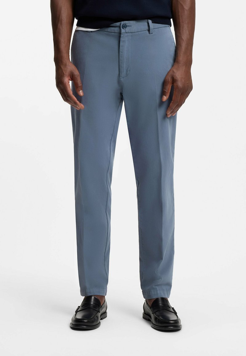 Homme portant un pantalon bleu sur mesure et des mocassins en cuir noirs, debout devant un fond blanc uni.