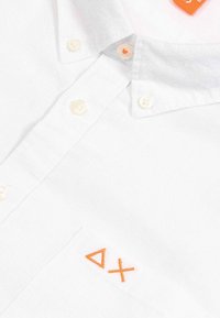 Camicia di cotone bianca con colletto corto, caratterizzata da simboli ricamati arancioni sulla tasca e bottoni in plastica. Tessuto testurizzato con una sottile trama.