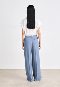Pantalons légers bleu clair, évasés à taille haute avec des plis, dotés d'un petit détail de poche marine à l'arrière. Associés à un haut blanc.