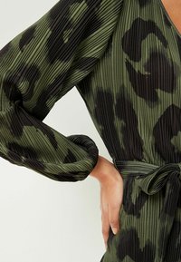 Vestido plisado verde con estampado de leopardo negro, con mangas largas y cintura ceñida con cinturón. Tejido texturizado con un brillo sutil.
