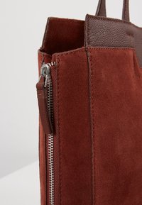 Sac fourre-tout en daim rouge avec des accents en cuir marron texturé, doté d'une fermeture éclair et de poignées robustes. Forme rectangulaire, fermeture éclair en métal lisse.