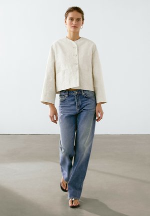Giacca bianca corta con scollo rotondo e chiusura a bottoni, abbinata a jeans azzurri dal taglio rilassato e sandali neri con dettagli metallici.