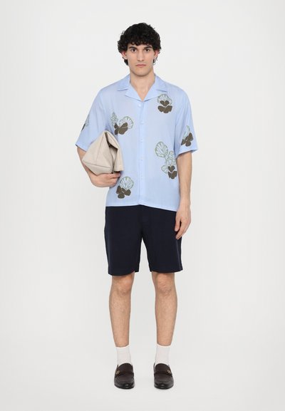Kortermet lyseblå skjorte med bladmønster, kombinert med mørke shorts. Holdt en beige veske, iført mørke slip-ons og hvite sokker.