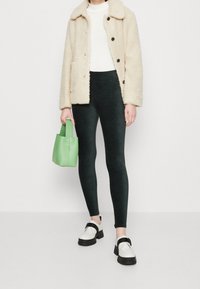 Persona che indossa una giacca in pile crema, una maglietta bianca, leggings neri e scarpe bianche spesse, mentre tiene una piccola borsa verde, in piedi di fronte a uno sfondo semplice.