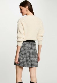 Cremesfarbener gerippter Pullover mit übergroßen Ärmeln, kombiniert mit einem Rock aus schwarz-weißem Tweed. Das Outfit beinhaltet einen schwarzen Gürtel. Glatte Texturen.