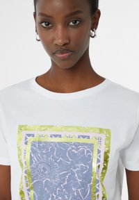 Witte katoenen t-shirt met een blauw bloemmotief omringd door metallic gele en roze randen. Ronde hals, korte mouwen, gladde textuur.