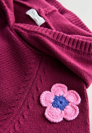 Magenta pleteni pulover s rebrastim ovratnikom, s detaljem kvačkanog cvijeta u ružičastoj i plavoj boji na prednjoj strani. Mekana tekstura i ležeran dizajn.