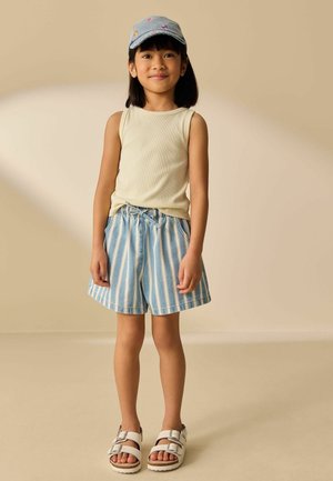Młoda dziewczyna w jasnym topie na ramiączkach, niebiesko-białych pasiastych szortach, białych sandałach i ozdobionej szarej czapce stojąca na beżowym tle.