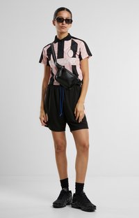 Polo majica v roza in črni barvi s šahovnato obrobo, v kombinaciji s črnimi kratkimi hlačami. Dodatki vključujejo malo črno torbico za čez ramo in sončna očala.