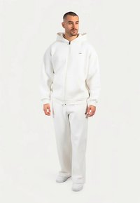 Weißer Kapuzenpullover mit Reißverschluss und Kordelzugkapuze, kombiniert mit passenden weißen, locker sitzenden Hosen und weißen Turnschuhen, die eine glatte Textur zeigen.