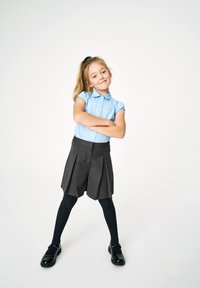 Camicia azzurra chiaro con colletto, pantaloni corti grigi plissettati, collant neri e scarpe in vernice. Il modello sta in piedi con sicurezza con le braccia incrociate.