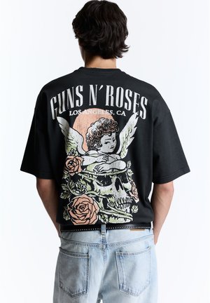 Személy fekete Guns N' Roses pólót visel angyal, koponya és rózsák mintával, világoskék farmernadrággal párosítva, hátulról látható.
