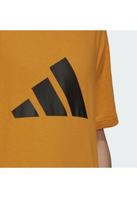 Senapsgul T-shirt i mjuk bomull, med en framträdande svart logotyp med tre ränder tryckt på bröstet.