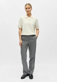 Kreemjas lühikeste varrukatega polo särgiga, millel on krae ja nööbid, koos hallide triibuliste pükstega. Modell seisab mustades kingades, ühtlane sile tekstuur kogu komplekti ulatuses.