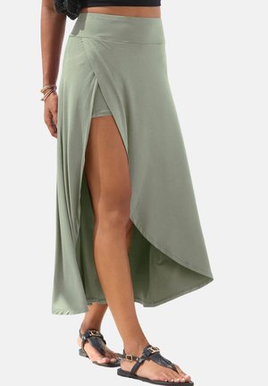 Frau trägt einen Wickelrock in Salbeigrün mit innerer Shorts, kombiniert mit schwarzen offenen Sandalen, gezeigt vom Bauch bis zu den Füßen.