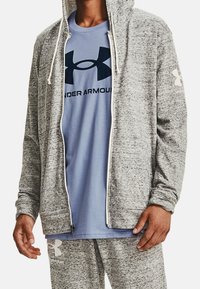 Felpa grigia con texture e zip, con lacci bianchi, indossata sopra una t-shirt azzurro chiaro con un logo scuro. Sono visibili anche pantaloni della tuta grigi.