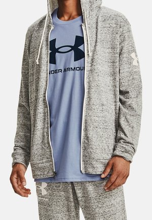 Sudadera con cremallera texturizada en gris y cordones blancos, llevada sobre una camiseta de color azul claro con un logo oscuro. También se ven pantalones de chándal grises.