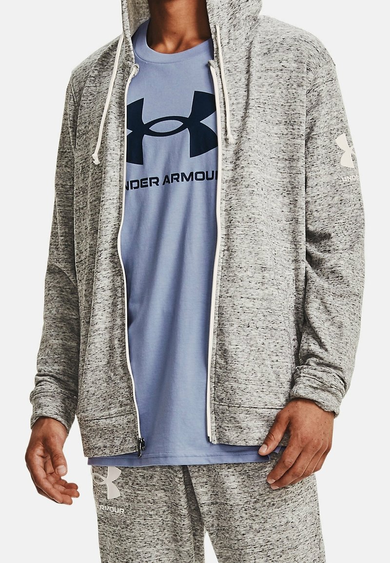 Felpa grigia con texture e zip, con lacci bianchi, indossata sopra una t-shirt azzurro chiaro con un logo scuro. Sono visibili anche pantaloni della tuta grigi.