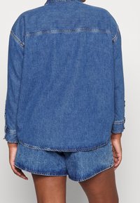 Henkilö pukeutuneena siniseen farkkutakkiin ja vastaaviin farkkushortseihin, näkyvissä takaa yksinkertaista taustaa vasten.