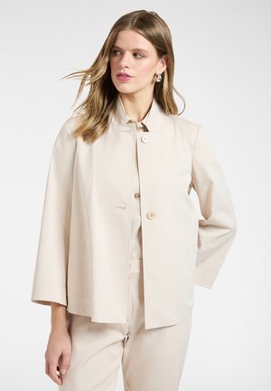 Giovane donna con capelli castano chiaro che indossa una giacca beige abbottonata e pantaloni abbinati, che guarda verso sinistra su uno sfondo bianco.