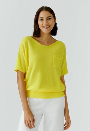 Frau mit schulterlangen dunklen Haaren, die lächelt, trägt ein hellgelbes kurzärmeliges Oberteil mit Brusttasche und weiße Hosen vor einem einfarbigen Hintergrund.