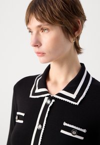 Chemise en tricot noir avec un col blanc à bords festonnés, des boutons et des rayures sur la poche. Présente une finition texturée et une coupe ajustée.