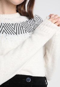 Vit stickad tröja med svart geometriskt mönster vid kragen, förlängda ärmar och ribbad detalj. Matchad med svarta kordbyxor.