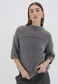 Szürke kötött pulóver magas gallérral, rövid ujjakkal és laza fazonnal. A texturált minta nyitott és zárt öltések keverékéből áll.