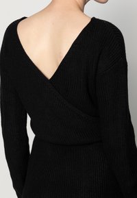 Pull en maille noire avec un dos en V profond. Doté de manches longues et d'un tissu texturé. Silhouette ajustée qui accentue la taille.