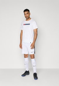 Hvid kortærmet T-shirt med et multikolor Nike-logo og teksten "PARIS SQ", parret med hvide shorts, hvide sokker og sorte fodboldsko.