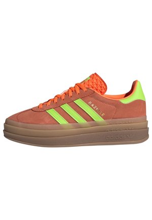 Baskets Adidas Gazelle en daim marron avec des lacets orange vif, des bandes vert néon et une languette au talon, ainsi qu'une semelle épaisse en caoutchouc marron.
