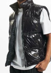 Gilet imbottito nero con texture lucida e riflettente, chiusura con zip e collo alto, indossato sopra una T-shirt bianca. Il gilet presenta tasche laterali.