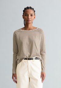 Frau mit geflochtenem Haar, die ein braun-weiß gestreiftes Langarmshirt trägt, das in eine hoch taillierte, cremefarbene Hose mit schwarzem Gürtel gesteckt ist.