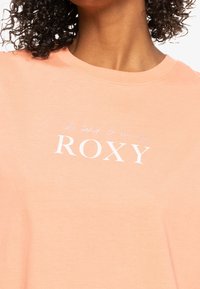 Krátký rukáv broskvové tričko vyrobené z měkké bavlny, s bílou grafikou "ROXY" a kurzívou, kulatým výstřihem a volnějším střihem.