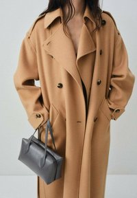 Camel trench coat with large lapels, double-breasted buttons, and side pockets. Accompanied by a small gray leather handbag.