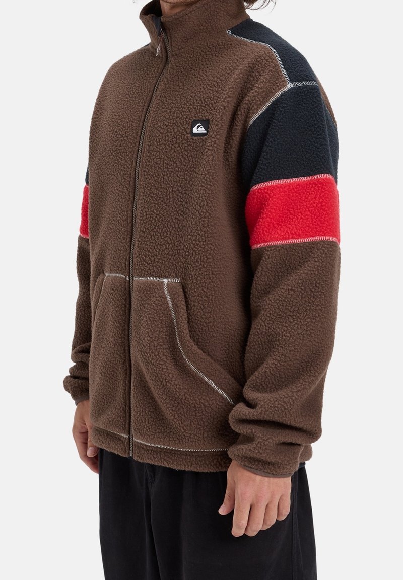 Homme portant une veste polaire marron avec des manches noires et rouges en bloc de couleurs, des poches avant et un petit logo sur la poitrine.