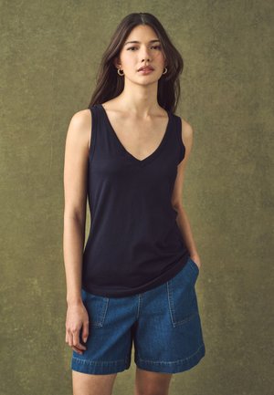 Jeune femme aux longs cheveux foncés portant des boucles d’oreilles créoles dorées, un haut noir sans manches à col en V et un short en jean bleu sur fond vert.