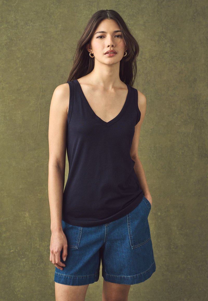 Jeune femme aux longs cheveux foncés portant des boucles d’oreilles créoles dorées, un haut noir sans manches à col en V et un short en jean bleu sur fond vert.