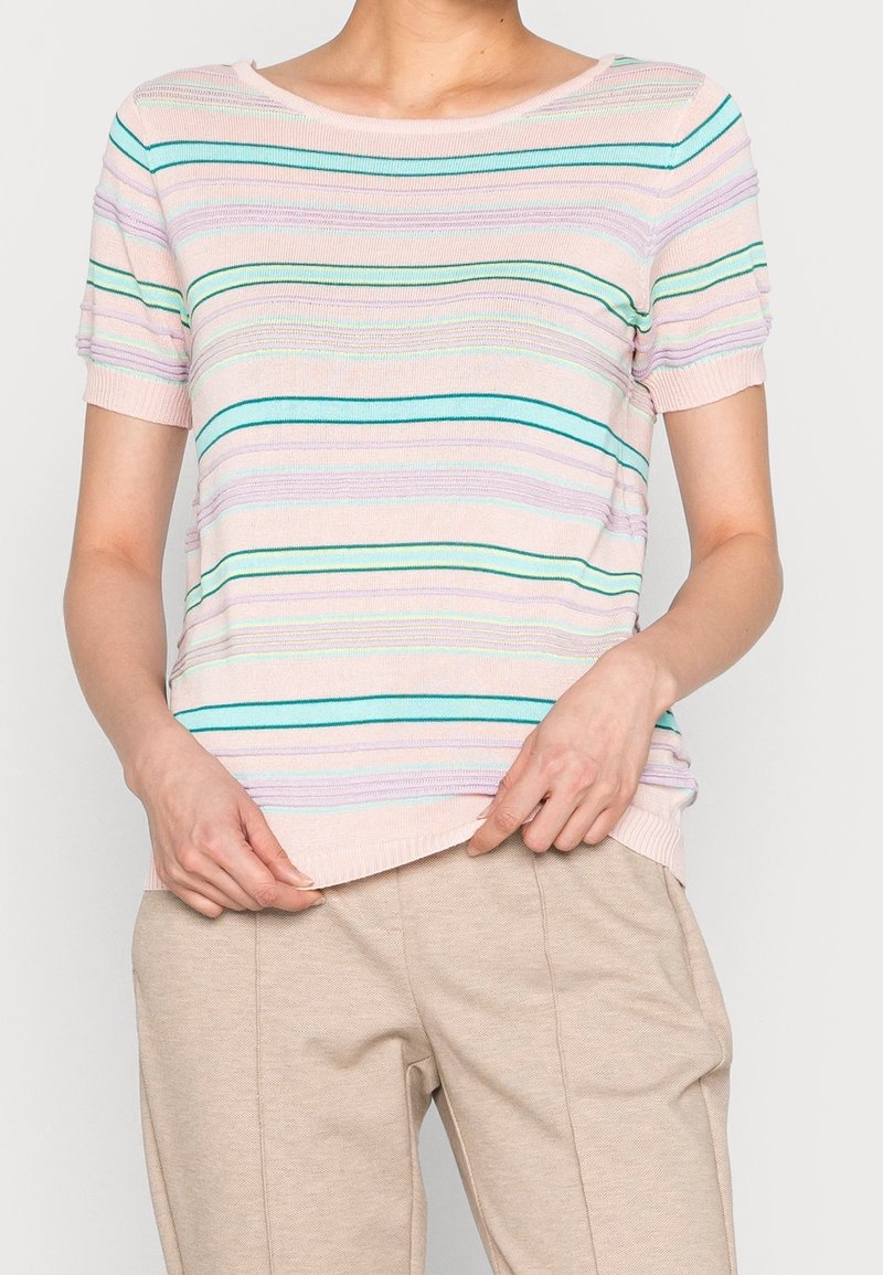 Femme portant un haut en maille rose pastel à manches courtes avec des rayures horizontales vertes et violettes, associé à un pantalon beige.