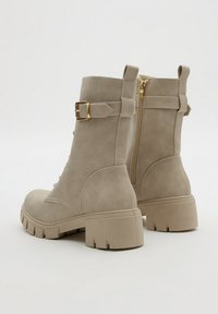 Botas de tobillo de gamuza topo con tacones gruesos, cremallera lateral y detalle decorativo de correa con hebilla dorada.