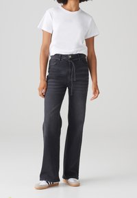 Personne portant un t-shirt blanc à manches courtes, un jean noir taille haute à jambes larges avec une ceinture tressée, et des baskets blanches avec des rayures foncées.