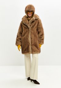 Capotă din blană sintetică maro, cu un guler mare și închidere frontală cu capse, asortată cu o pălărie maro, mănuși galbene și pantaloni largi crem.