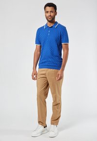 Sininen teksturoitu polo-paita, jossa on ribbireuna kauluksessa ja helmassa, yhdistettynä vaaleanruskeisiin housuihin ja valkoisiin lenkkitossuihin.