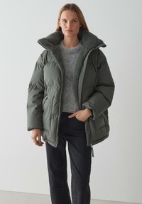 Giacca imbottita verde oliva con un ampio collo morbido e testurizzato, chiusura con zip e tasche laterali, abbinata a un maglione grigio e jeans scuri.