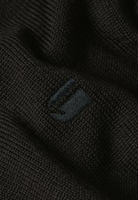 Schwarzer Strickstoff mit strukturierter Oberfläche; verfügt über ein kleines, blaues Stofflogo, das auf das Material gestickt ist.