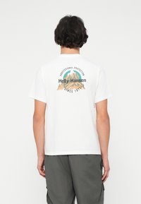 Mladic s kratkom tamnom kosom nosi bijelu majicu s planinskim logotipom i natpisom "Helly-Hansen Profesionalna zaštita od 1877." na leđima.