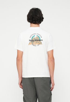 Jovem com cabelo curto e escuro, a usar uma t-shirt branca com um logótipo de montanha e o texto "Helly-Hansen Professional Protection Since 1877" nas costas.