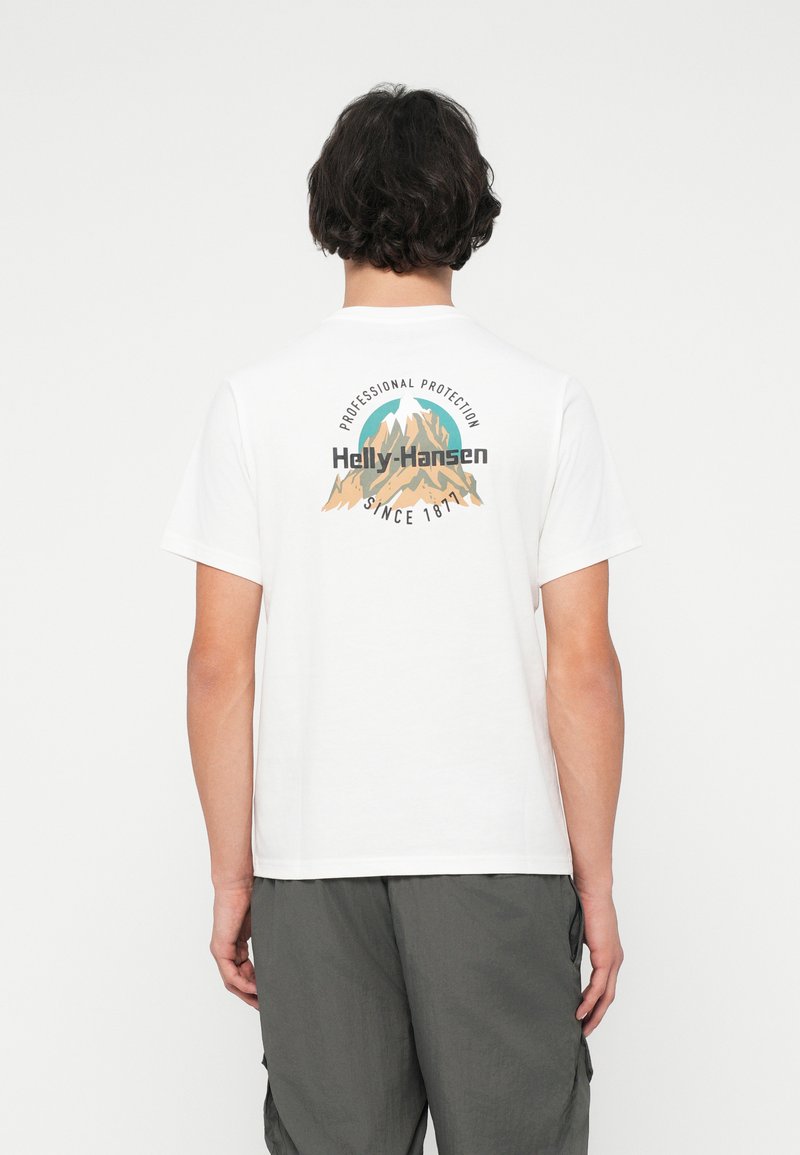 Jovem com cabelo curto e escuro, a usar uma t-shirt branca com um logótipo de montanha e o texto "Helly-Hansen Professional Protection Since 1877" nas costas.